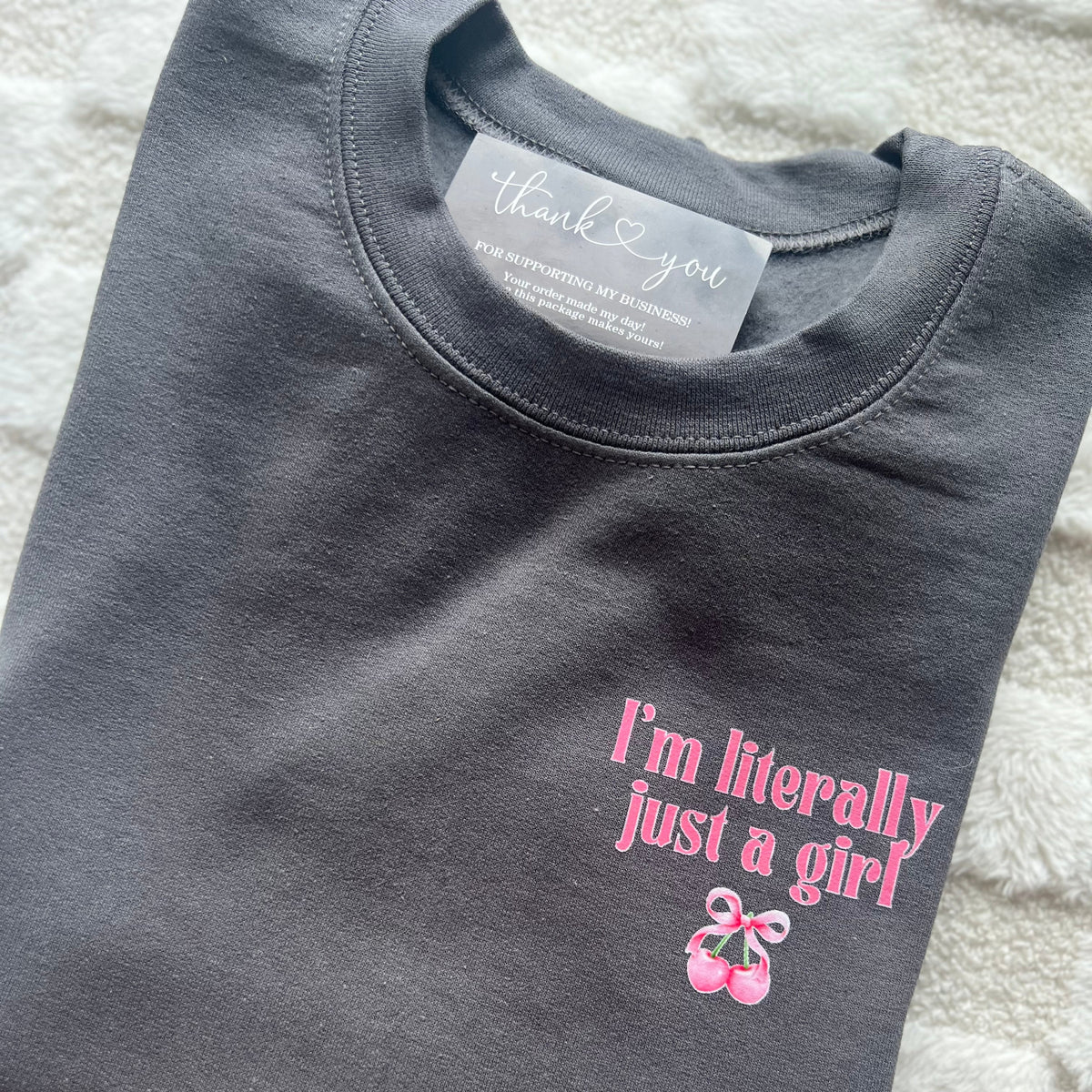 A gray crewneck sweatshirt with the text 'I'm literally just a girl' printed in pink, accompanied by a small pink graphic of a bow & cherries.